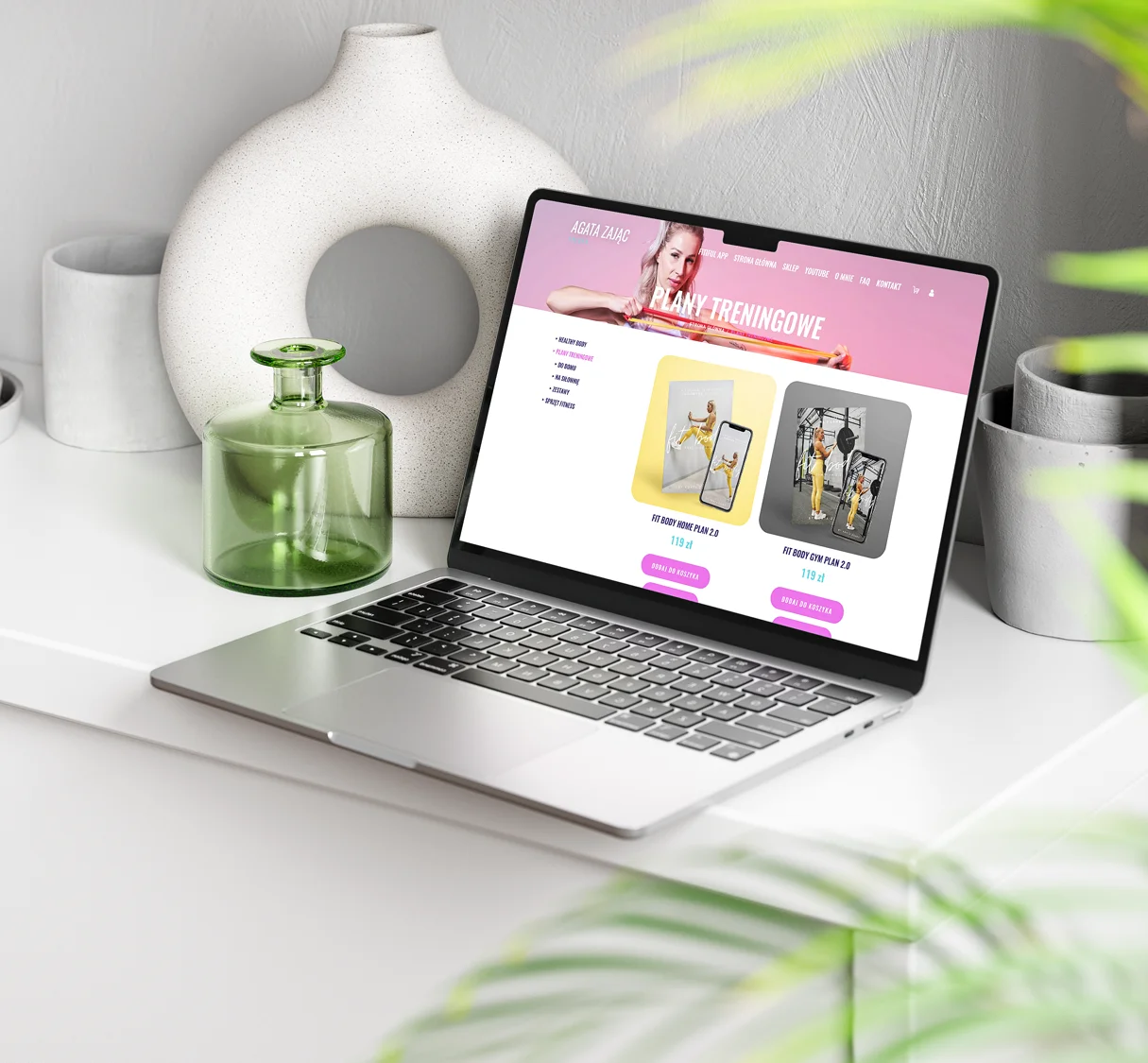 Obraz przedstawia laptopa na eleganckiej, nowoczesnej toaletce, na którym otwarta jest strona internetowa oferująca plany treningowe. Wokół laptopa znajdują się stylowe dekoracje, takie jak szklane wazony i ceramiczne elementy, co nadaje estetyczny i uporządkowany wygląd