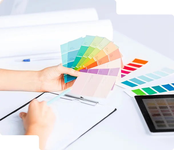 Obraz przedstawia proces wyboru kolorów w projektowaniu graficznym lub brandingowym. Osoba trzyma wachlarz próbek kolorystycznych, prawdopodobnie z palety Pantone lub RAL